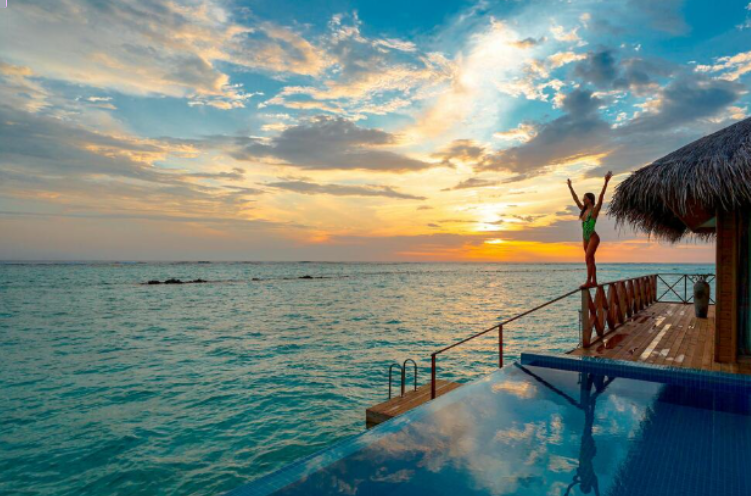 A traveler celebrates sunset views from a stunning overwater villa in paradise.
