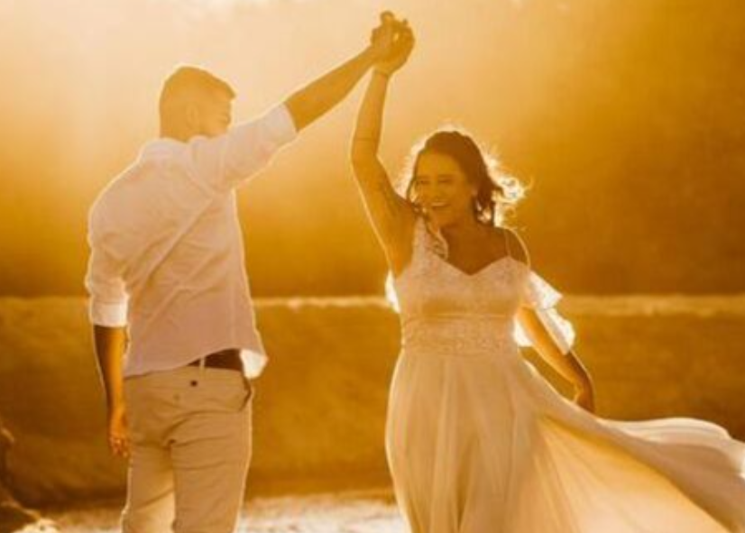 A newlywed couple dances joyfully by the water at golden hour, celebrating love in a breathtaking sunset setting.