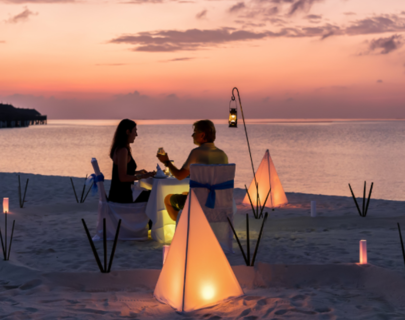 A couple enjoys an intimate candlelit dinner on the beach at sunset, surrounded by soft glowing lights and ocean views.
