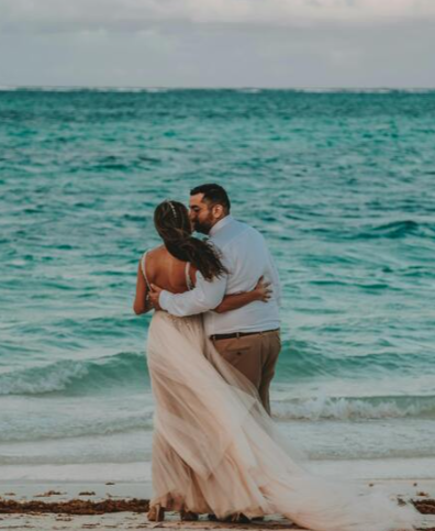 Newlyweds embrace by the shoreline, sharing a quiet moment as waves gently meet the sand.