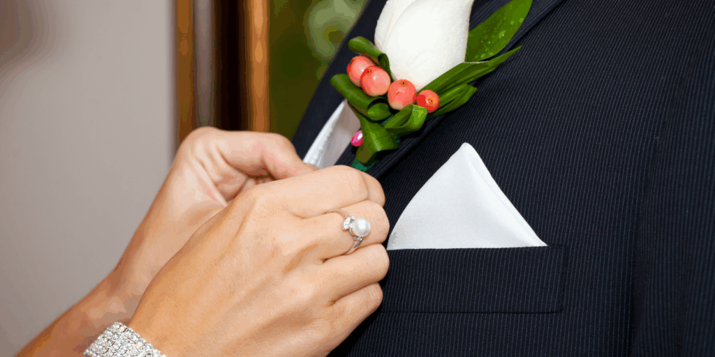 Getting-ready details before the ceremony begins, a woman pins a boutonniere on the lapel of wedding party member.