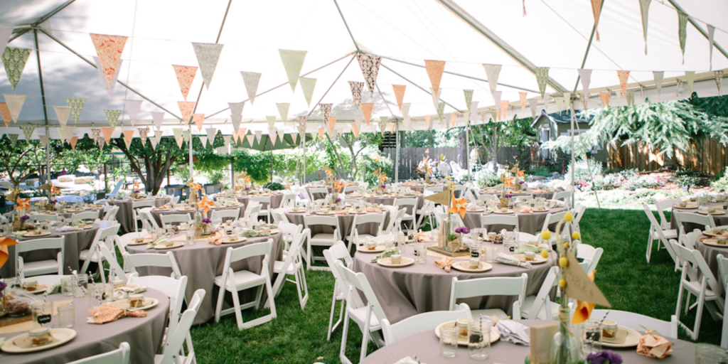 Wedding reception setup under a tent on a lawn, with round tables, white chairs, and decorative bunting.