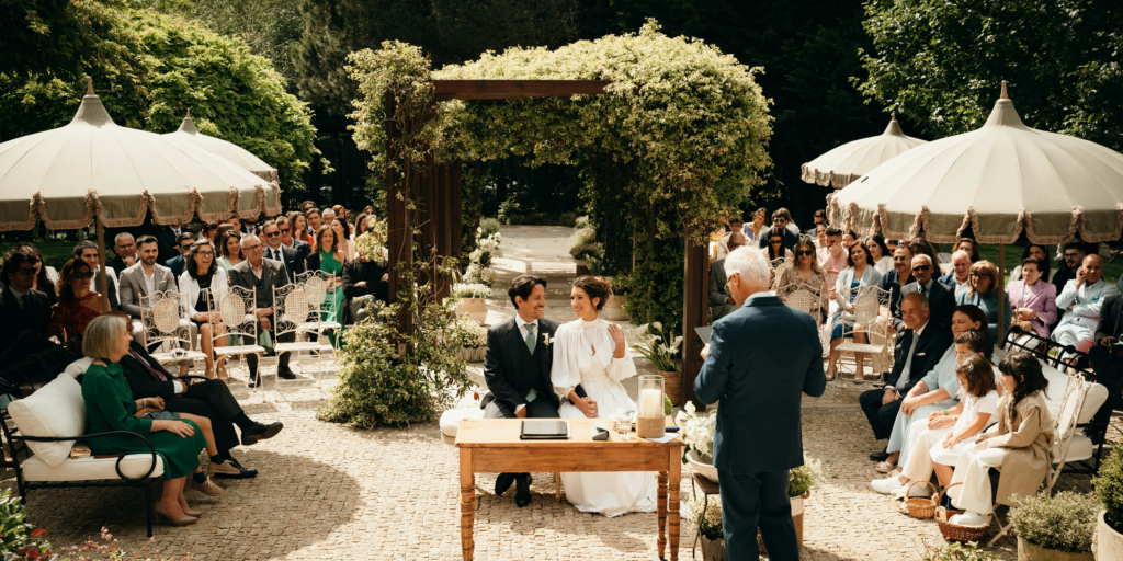 Elegant outdoor wedding ceremony with bride and groom seated under greenery arch surrounded by guests in garden setting.