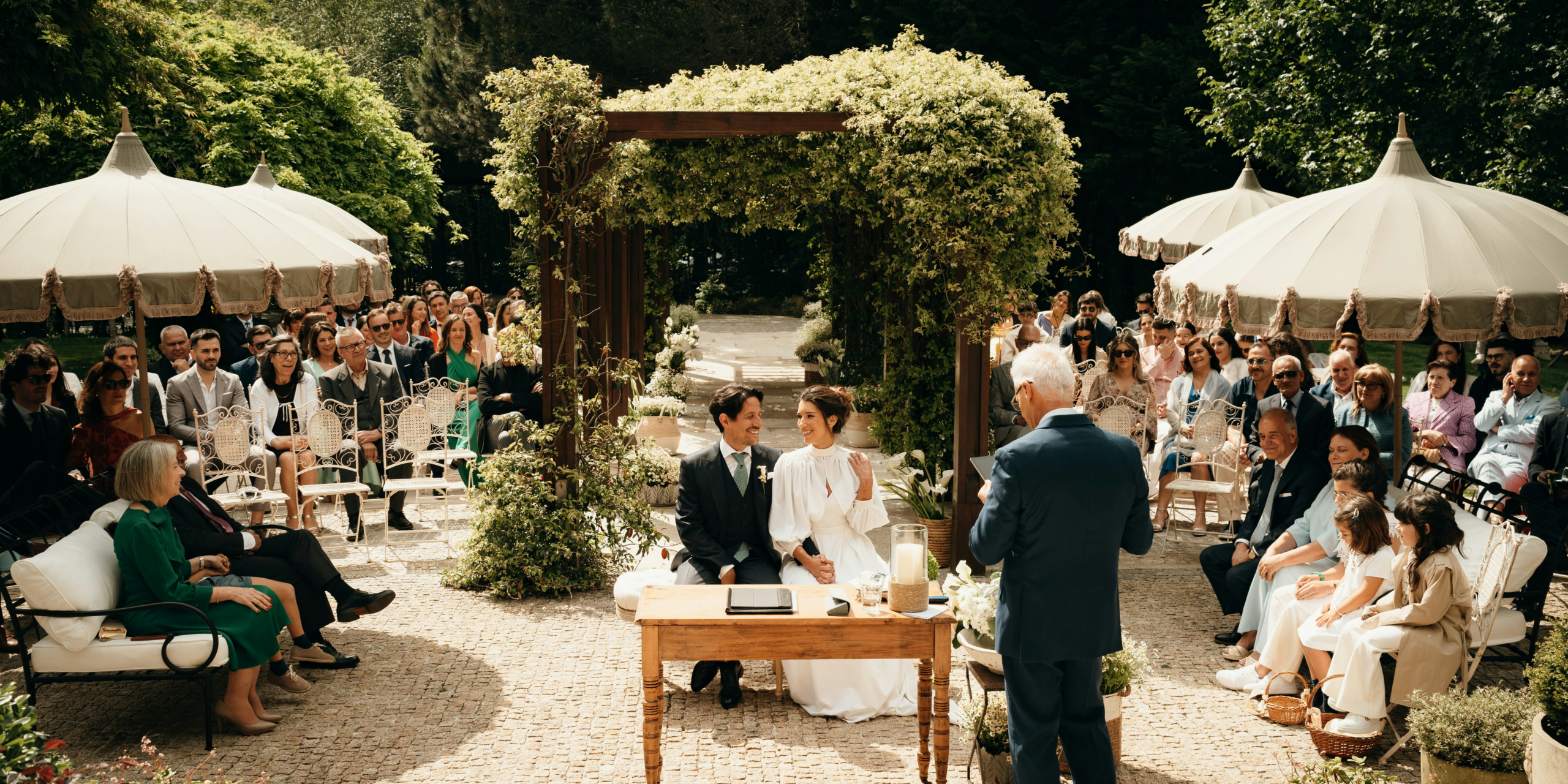 Elegant outdoor wedding ceremony with bride and groom seated under greenery arch surrounded by guests in garden setting.