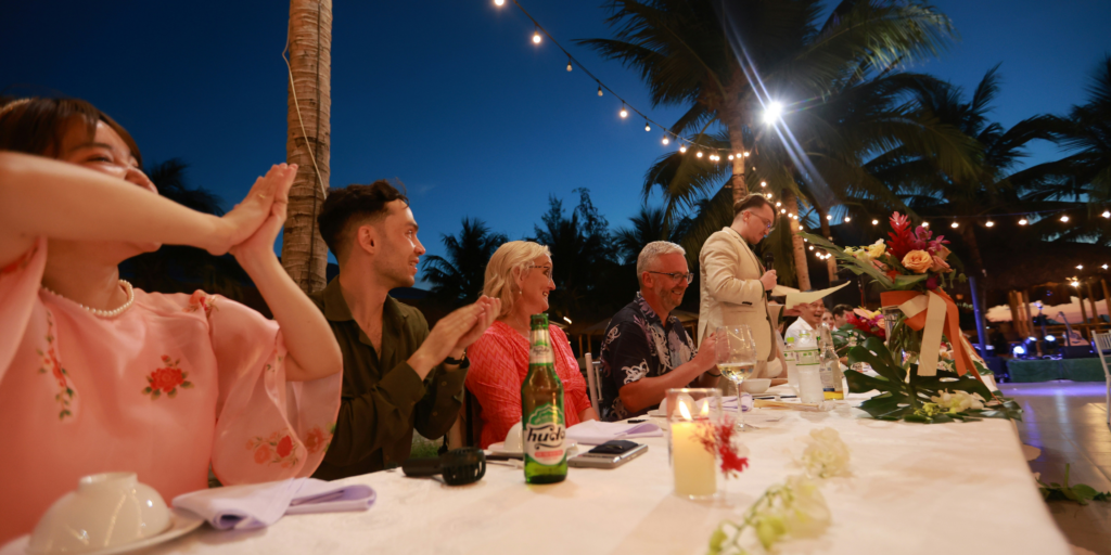 Evening wedding reception with family applauding speech under string lights and palm trees.