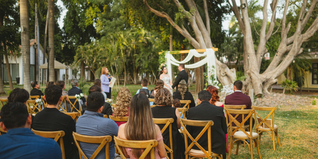 Garden wedding ceremony with guests seated on wooden chairs facing floral arch.