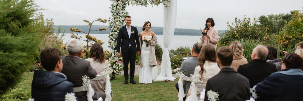 Intimate outdoor wedding ceremony with the couple standing beneath a floral arch as close family and friends sit nearby overlooking a lake.